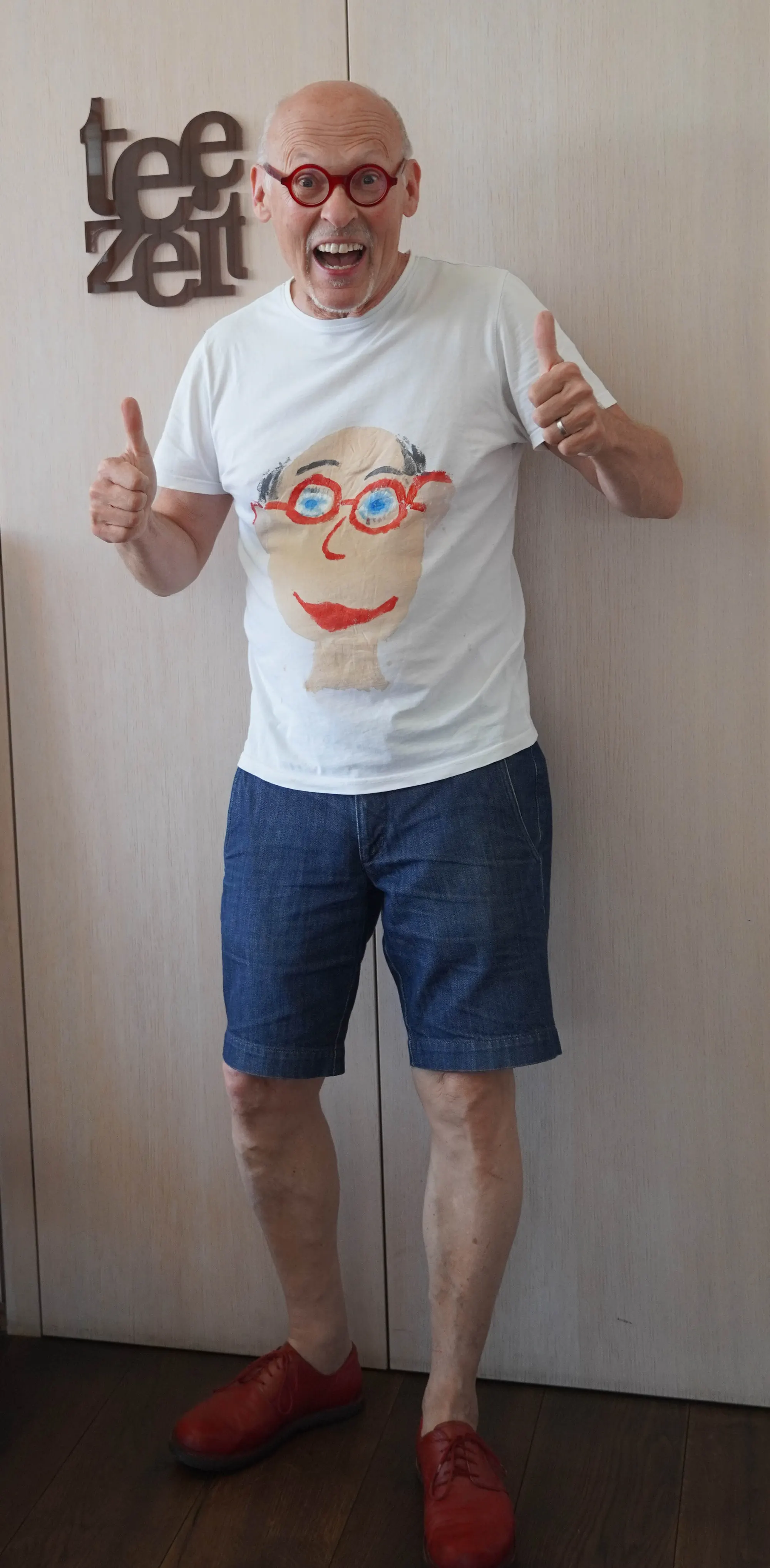 Johannes Gutmann en pied, lunettes rondes rouges, pouces levés, sous l’inscription « Teezeit »