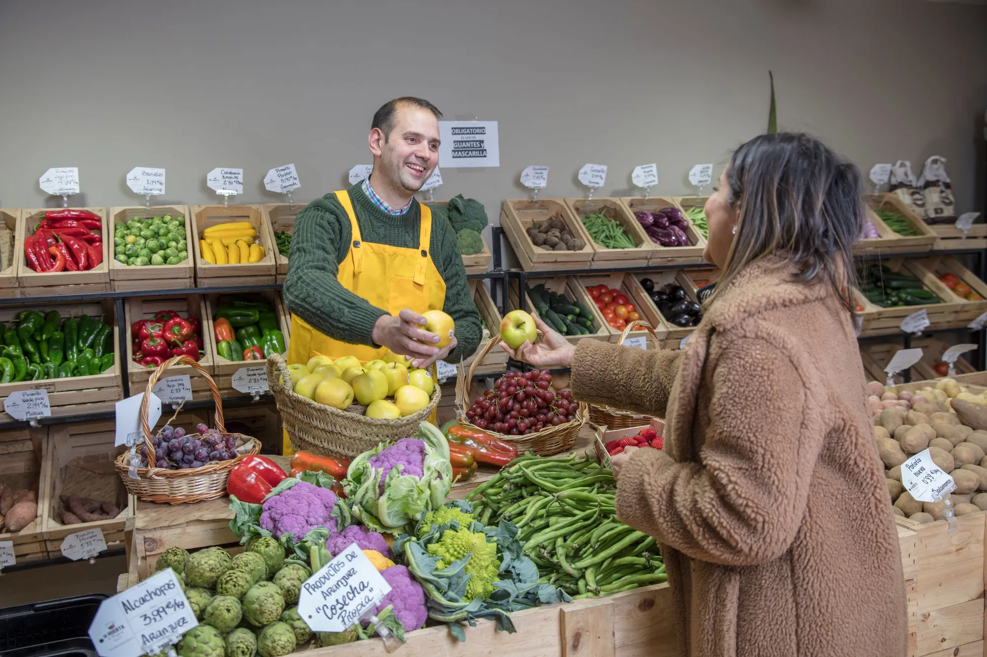 Un primeur tend une pomme à une cliente, devant ses cagettes de légumes étiquetées à la main