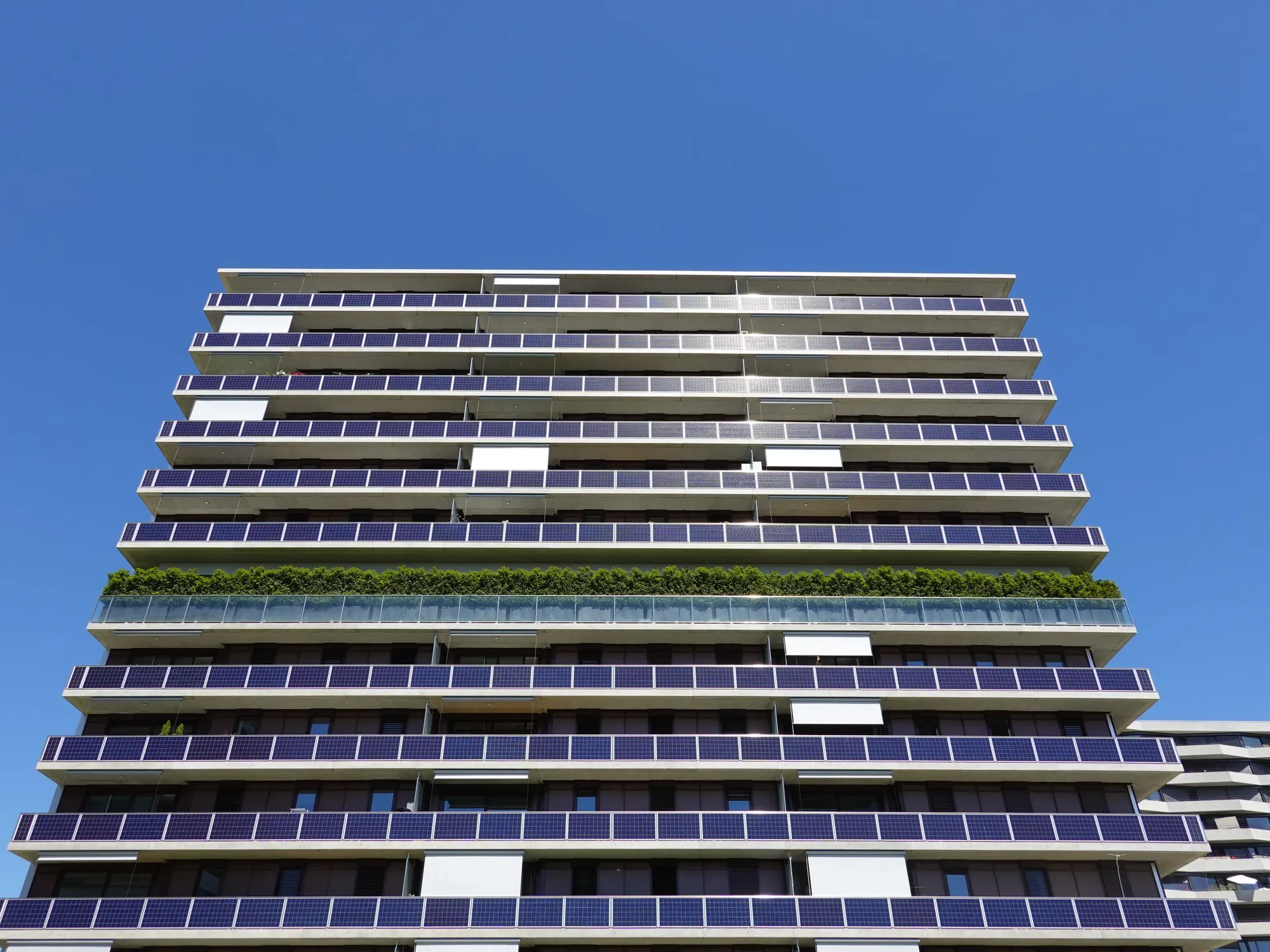 Façade de la tour des Vergers en contre-plongée : garde-corps couverts de panneaux solaires, étage végétalisé, ciel bleu, juin 2020