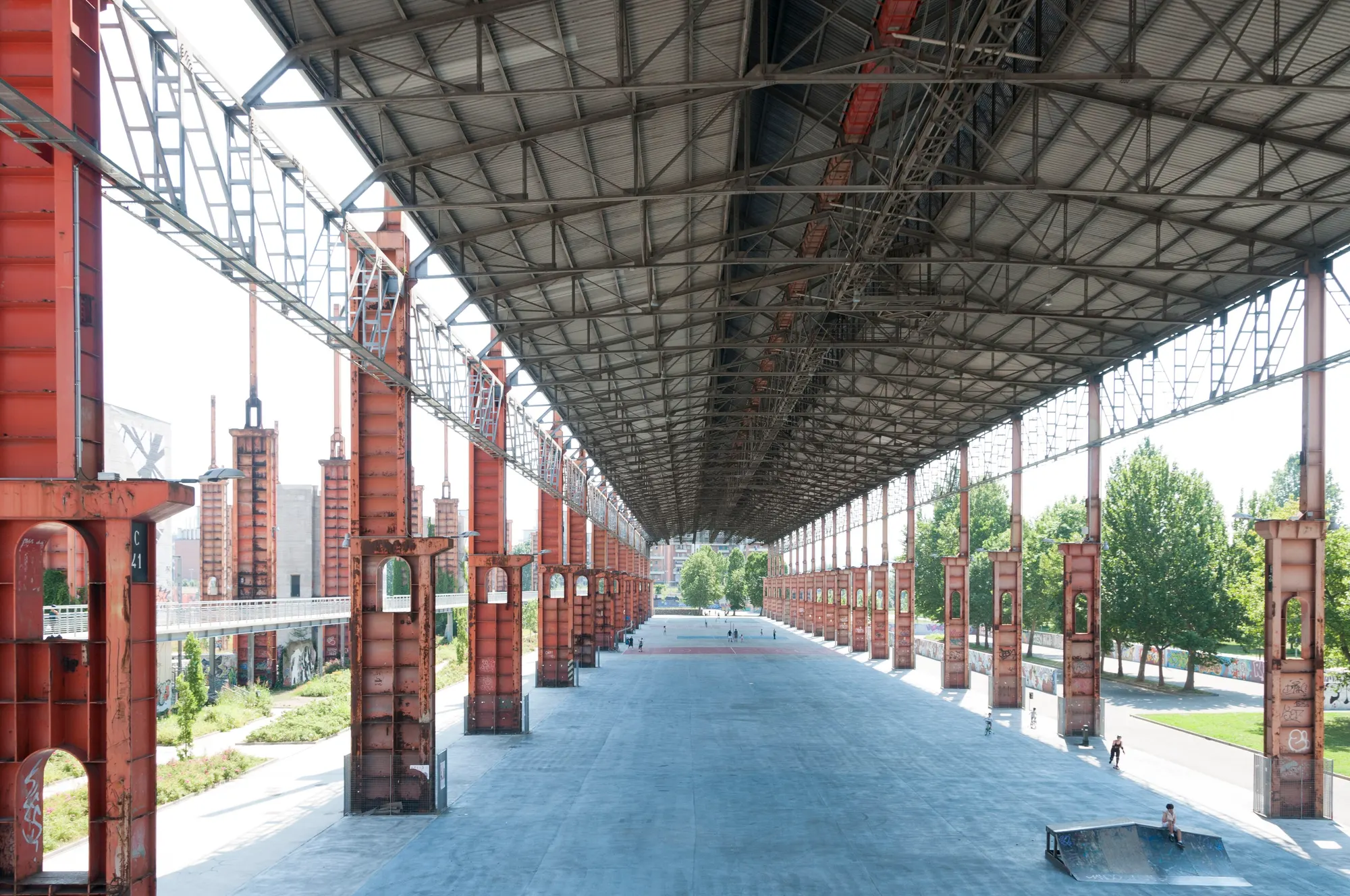 Sous la halle du strippaggio : charpente métallique, piliers rouges en enfilade, skatepark, enfants à vélo, juin 2015