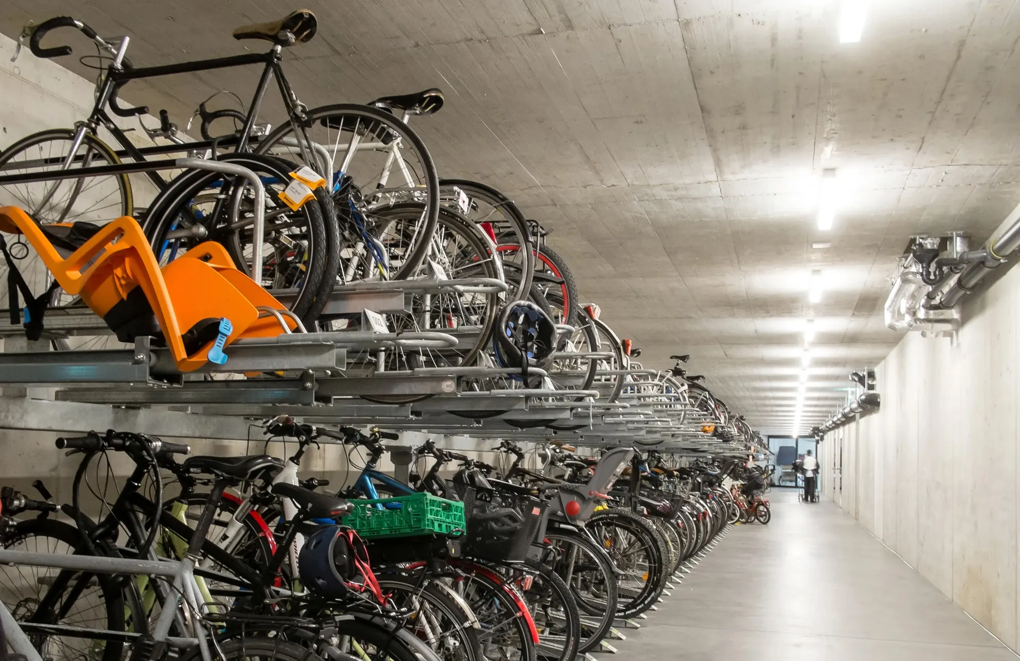 Le garage à vélos : râteliers à deux niveaux pleins, siège enfant orange, long couloir de béton, 2016
