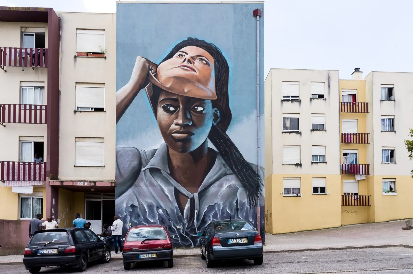 Fresque de Nomen sur un pignon : femme noire qui retire un masque blanc, immeubles de logement social et voitures garées au pied, juin 2016